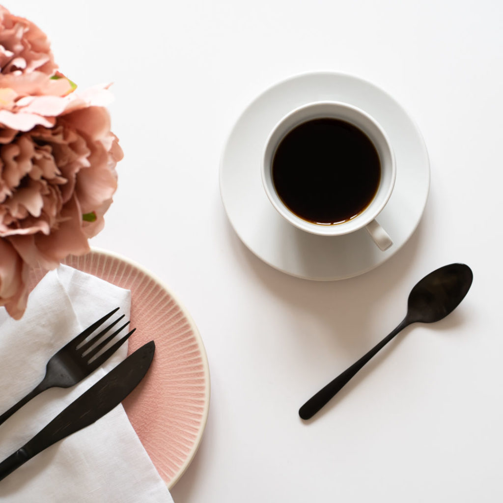 Flatlay s kávou v bílém šálku, s růžovým talířkem a černými příbory a růžovými pivoňkami